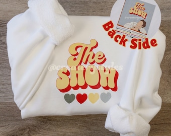 Puede incluir: Sudadera blanca con el texto "The Show" en fuente de estilo retro con acentos amarillos, rojos y grises. Hay cuatro pequeños corazones en gris, rojo y amarillo debajo del texto. La parte posterior de la sudadera tiene una pegatina con el texto "The Show" y una imagen de una persona.