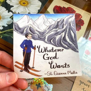 May include: A square sticker featuring a watercolor illustration of a person skiing down a snowy mountain. The person wears a blue sweater and black pants. The sticker has the text "Whatever God Wants ~St. Gianna Molla."