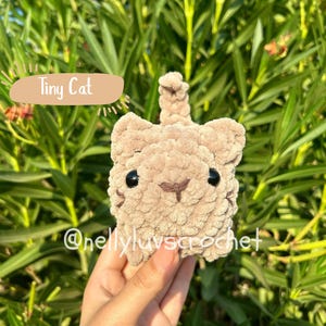 May include: A crocheted tiny cat in a light brown colour with black eyes and a small brown nose. The cat has a small tail and the text "Tiny Cat" is on a light brown banner. The text "@nellyluvscrochet" is also visible.