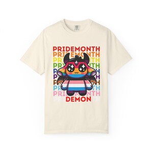 May include: A cream-coloured t-shirt with a cartoon demon character wearing sunglasses and a transgender flag design. The text "PRIDEMONTH" is stacked vertically in rainbow colours, with "DEMON" at the bottom.