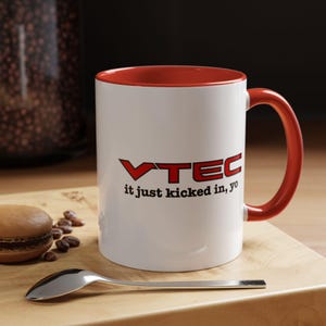 May include: A white ceramic mug with a red interior and a red handle. The front of the mug features the red text VTEC and the black text it just kicked in, yo. The mug is positioned on a wooden surface next to a silver spoon and a brown macaron.