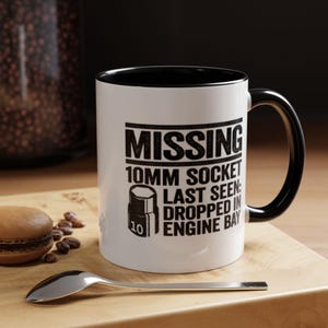 May include: A white ceramic mug with a black interior and handle. The front features black text that reads MISSING 10MM SOCKET LAST SEEN: DROPPED IN ENGINE BAY, accompanied by a graphic illustration of a socket labeled with the number 10.