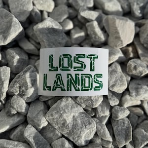 May include: A close-up of a pile of small, gray rocks with a white sticker that says "LOST LANDS" in green letters.