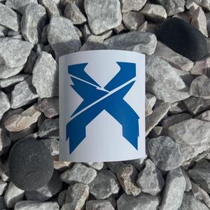 May include: A white sticker with a blue geometric design in the shape of an X. The sticker is placed on a bed of gray and white rocks, with two black stones. The design is a bold, modern graphic.