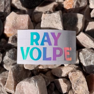 May include: A white sticker with the name "RAY VOLPE" in a gradient of colors, including teal, purple, and orange. The sticker is placed on a bed of small, light-colored rocks.
