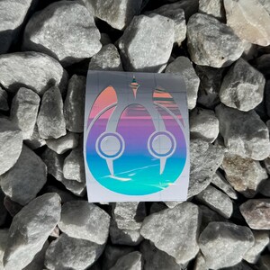 May include: A holographic sticker with a stylized design featuring a central structure with two circular elements and pointed accents. The sticker transitions from blue to purple and orange hues. The sticker is placed on a bed of gray rocks.