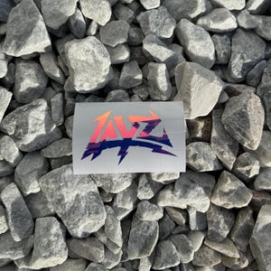 May include: A white sticker with a holographic finish featuring the word "TAVZ" in a stylized font with a lightning bolt underneath. The letters are in a gradient of pink, orange, and blue.