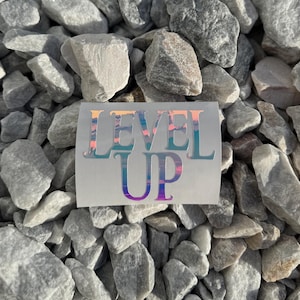 May include: A sticker with the words "LEVEL UP" in a holographic font with a rainbow color scheme. The sticker is on a gray background of small rocks.