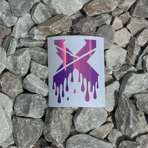 May include: A white sticker featuring a stylized, dripping letter "X" in shades of pink and purple. The design has a metallic, iridescent finish. The sticker is set against a background of gray rocks.