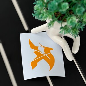 May include: A white sticker with a bright orange stylized bird design. The sticker is on a black surface.