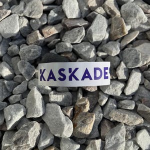 May include: Close-up of a white label with the word "KASKADE" printed in purple letters on a bed of gray gravel.