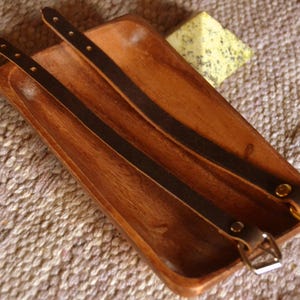 Op de afbeelding: Twee bruine leren riemen met gouden metalen gespen op een houten dienblad.