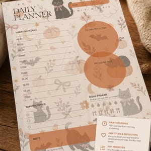 May include: A daily planner sheet featuring a cream background with illustrations of black cats, ghosts, pumpkins, and flowers. The layout includes sections for a schedule, gratitude, goals, and priorities, with text labels like Daily Planner and Note.