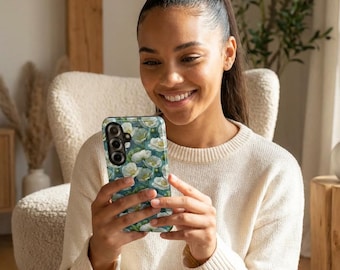 May include: A floral phone case featuring a pattern of white flowers on a teal background. The case has a camera cutout and button placements. The phone case is held by a person wearing a cream-colored sweater.