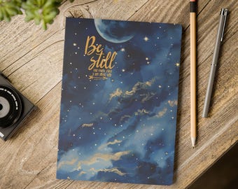 May include: A notebook with a celestial design featuring a navy blue background, a crescent moon, and gold stars and clouds. The cover has the words "Be still" in gold script. A pencil and pen are next to the notebook.