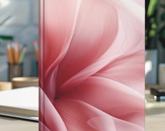 May include: A hardcover notebook with a pink floral design. The cover features a close-up of soft, flowing petals in shades of pink and white, creating a delicate and artistic aesthetic. The notebook is standing upright on a light-colored surface.