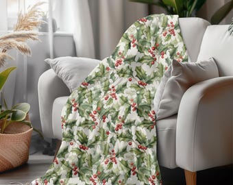 May include: A cozy blanket draped over a white sofa. The blanket features a repeating pattern of green holly leaves and red berries on a cream background. The blanket adds a festive touch to the living room setting.