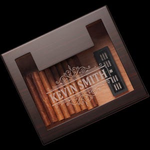 May include: A dark brown wooden cigar box with a glass top. The box is engraved with the name "KEVIN SMITH" and has a humidifier inside. The box is filled with cigars.