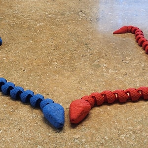 May include: Two flexible snake toys, one blue and one red, are posed facing each other on a brown surface. The snakes are made of segmented plastic and have a realistic snake head shape.