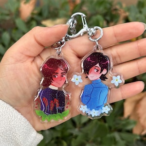 Puede incluir: Dos encantos de personajes en una cadena plateada. Los encantos son transparentes y presentan ilustraciones de dos personas. Un encanto tiene una persona con cabello castaño y una camisa azul, rodeada de flores azules. El otro encanto tiene una persona con cabello castaño y una camisa morada, rodeada de flores rojas.