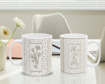 May include: Two white ceramic mugs with a floral design. The mugs have a white background with a beige floral pattern and a beige geometric pattern. The mugs are on a white surface.