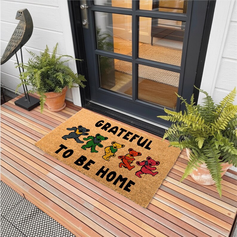May include: A rectangular doormat made of natural fibers. The mat features the text "GRATEFUL TO BE HOME" with dancing bear graphics in various colors. The doormat is placed on a wooden porch.