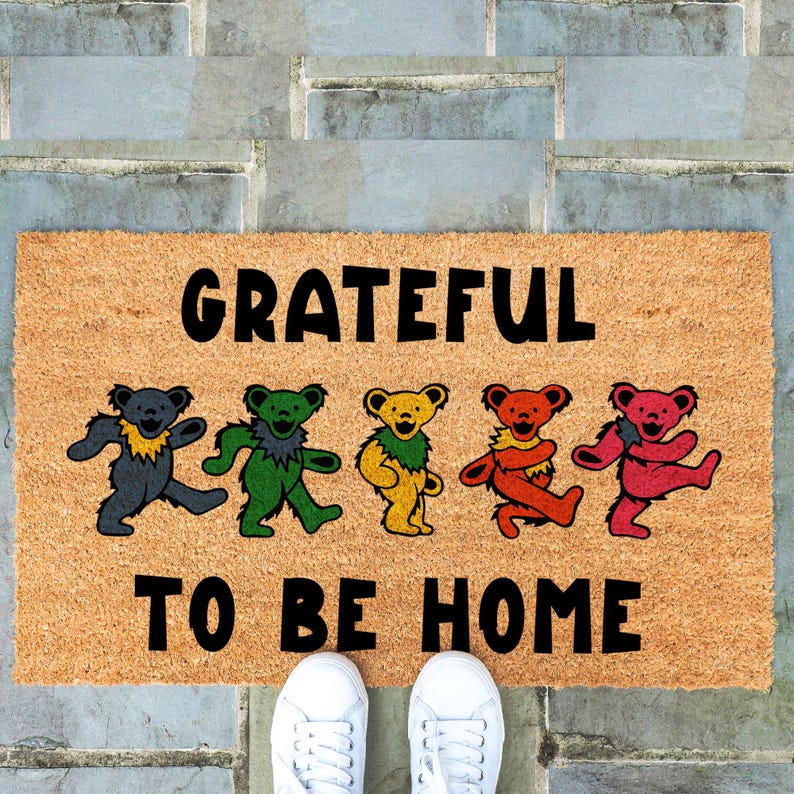 May include: A rectangular doormat with the words "GRATEFUL TO BE HOME" in black. It features dancing bear illustrations in various colors: gray, green, yellow, orange, and red. White sneakers are visible at the bottom.