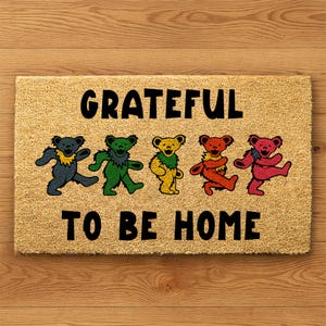 May include: Rectangular doormat with the words "GRATEFUL TO BE HOME" in black. Five colorful dancing bears in blue, green, yellow, orange, and pink are featured below the word "GRATEFUL". The doormat is made of a tan, natural fiber material.