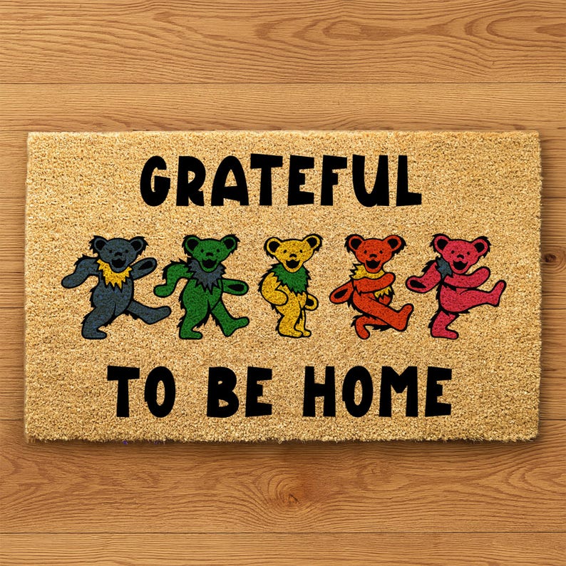 May include: Rectangular doormat with the words "GRATEFUL TO BE HOME" in black. Five colorful dancing bears in blue, green, yellow, orange, and pink are featured below the word "GRATEFUL". The doormat is made of a tan, natural fiber material.