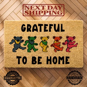 May include: A rectangular doormat made of natural fibers. The mat features the words "GRATEFUL TO BE HOME" in black block letters, with colorful dancing bear illustrations. The mat also has a "Satisfaction Guarantee" and "Handmade" seal.