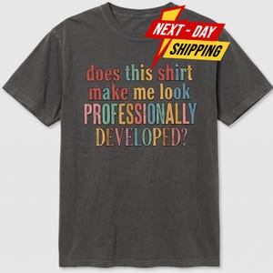 May include: A charcoal grey short-sleeve t-shirt featuring the multi-coloured text "does this shirt make me look PROFESSIONALLY DEVELOPED?" in a distressed, retro-inspired font. The shirt has a crew neckline and a standard, casual fit.