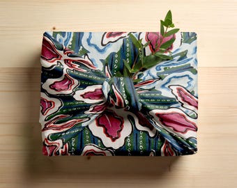 Può includere: Un regalo quadrato avvolto in un tessuto fantasia con un motivo botanico nei toni del blu, verde e rosa. Il tessuto è annodato e un rametto di verde è infilato nel nodo. Il regalo è su una superficie di legno chiaro.