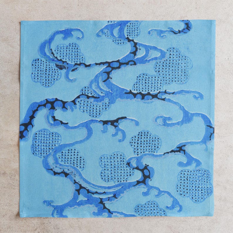 Pu&ograve; includere: Un tessuto quadrato azzurro con un motivo ripetuto di onde blu stilizzate e forme simili a nuvole. Le linee ondulate sono delineate in nero, creando un contrasto con lo sfondo azzurro pi&ugrave; chiaro. Le forme delle nuvole hanno una trama a pois.