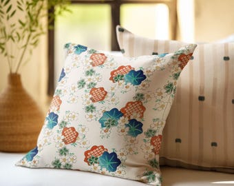 Könnte beinhalten: Ein weißes Kissen mit einem Blumenmuster mit blauen, grünen und orangefarbenen Blumen. Das Kissen liegt auf einem weißen Sofa mit einem gestreiften Kissen dahinter.
