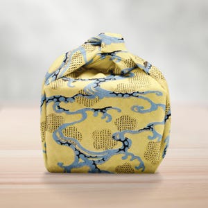 Pu&ograve; includere: Una borsa di stoffa gialla con un motivo a nuvole blu e nere. La borsa &egrave; annodata in alto, creando una forma unica. Il tessuto ha un aspetto strutturato.
