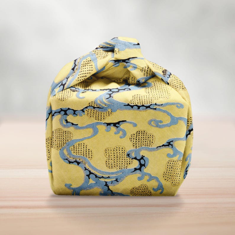 Pu&ograve; includere: Una borsa di stoffa gialla con un motivo a nuvole blu e nere. La borsa &egrave; annodata in alto, creando una forma unica. Il tessuto ha un aspetto strutturato.
