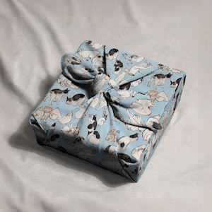 Pu&ograve; includere: Un pacco quadrato avvolto in tessuto azzurro, decorato con varie illustrazioni di gatti in nero, bianco e grigio. Il tessuto &egrave; legato in alto con un nodo, creando una chiusura decorativa. Il pacco &egrave; appoggiato su una superficie bianca.