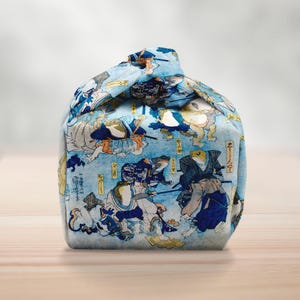 May include: A blue fabric bag with a Japanese woodblock print depicting frogs in samurai clothing. The bag is rounded and tied at the top. The print features blue, white, and yellow hues, creating a detailed pattern.