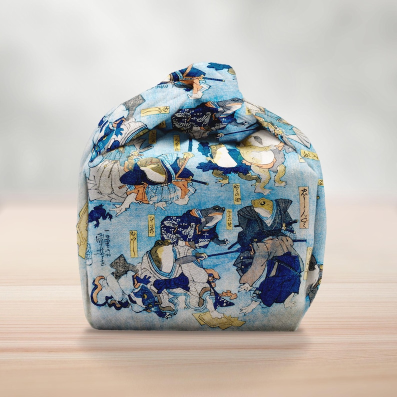 May include: A blue fabric bag with a Japanese woodblock print depicting frogs in samurai clothing. The bag is rounded and tied at the top. The print features blue, white, and yellow hues, creating a detailed pattern.