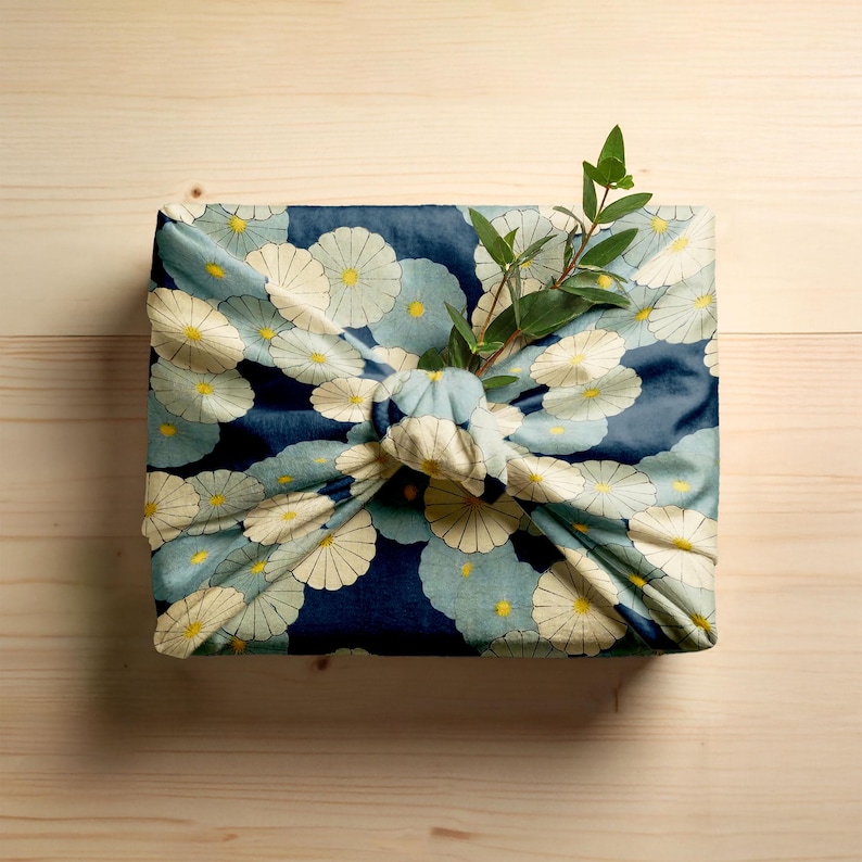 Gift box wrapped in Japanese Furoshiki style using floral fabric with cream and blue flowers on a navy background. Finished with a knotted top and a green leaf sprig for an elegant, eco-friendly presentation.