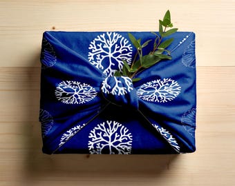 May include: A square, navy blue fabric wrapping with a white tree branch pattern. The fabric is tied in a knot, with a sprig of green leaves tucked into the knot. The wrapping is on a light wood surface.