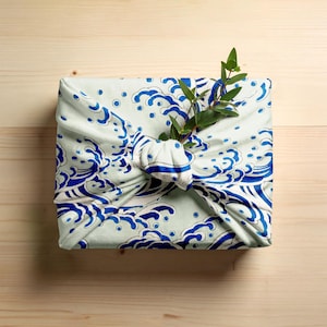 May include: A square package wrapped in a light green cloth with a blue wave pattern. A sprig of green leaves is tucked into the knot. The package is on a light wood surface.