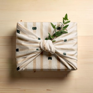 May include: A wrapped gift, presented in a beige and white striped fabric with dark green accents. The fabric is tied in a knot and adorned with a sprig of green leaves. The gift is placed on a light wooden surface.