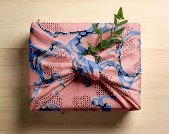 Può includere: Un panno di imballaggio in tessuto rosa con un motivo blu e nero, annodato. Un rametto di foglie verdi è infilato nel nodo. Il panno è su una superficie di legno chiaro.