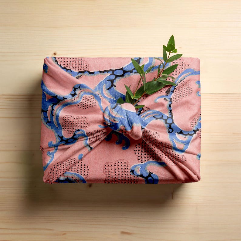 Pu&ograve; includere: Un panno di imballaggio in tessuto rosa con un motivo blu e nero, annodato. Un rametto di foglie verdi &egrave; infilato nel nodo. Il panno &egrave; su una superficie di legno chiaro.
