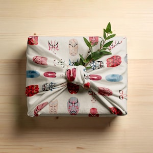 May include: A wrapped gift with a white cloth featuring a repeating pattern of Japanese Noh masks in red, pink, blue, and gray. A sprig of green leaves is tucked into the knot of the wrapping. The background is a light wood surface.