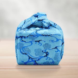 Pu&ograve; includere: Una borsa di stoffa blu con un motivo a nuvole e onde. La borsa &egrave; annodata in alto, creando una forma unica. Il design presenta uno sfondo azzurro con accenti blu scuro e neri.