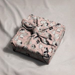 Pu&ograve; includere: Un regalo quadrato avvolto in tessuto rosa con un motivo ripetuto di illustrazioni di gatti in bianco e nero. Il tessuto &egrave; legato con un fiocco abbinato, creando una presentazione decorativa e unica.