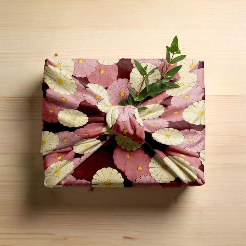 Pode incluir: Um pacote quadrado embrulhado em tecido com um padr&atilde;o floral. O tecido apresenta flores rosa e creme com centros amarelos sobre um fundo vermelho escuro. Um raminho de folhas verdes &eacute; colocado no n&oacute;.