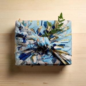 May include: A square package wrapped in blue fabric with a Japanese illustration, tied with a knot. The fabric depicts figures in blue, white, and tan. A sprig of green leaves is tucked into the knot.